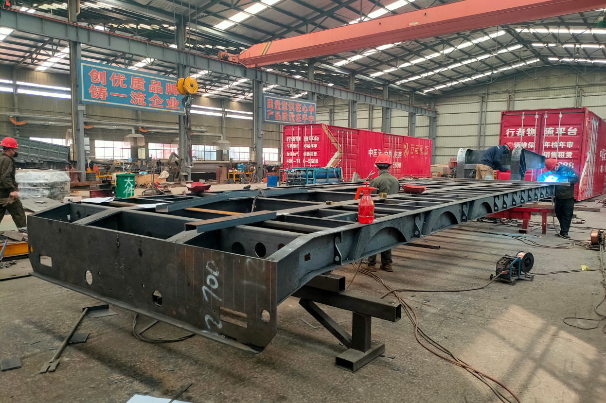 Factory workers performing precision welding on a heavy-duty lowbed trailer chassis, reinforcing the high-density cross-members for mining transport.
