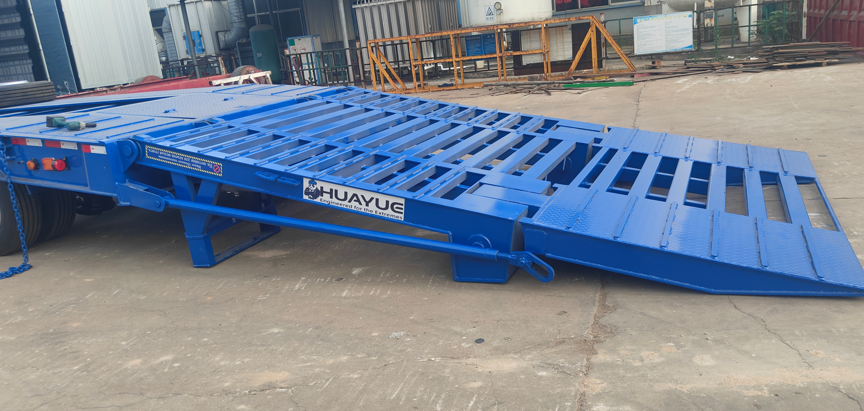 Side close-up of the HUAYUE lowbed trailer's double-folding hydraulic ramps. Clearly displaying the heavy-duty hydraulic cylinder support structure and the extremely low climbing angle formed when fully deployed, effectively preventing the seesaw effect during excavator loading.