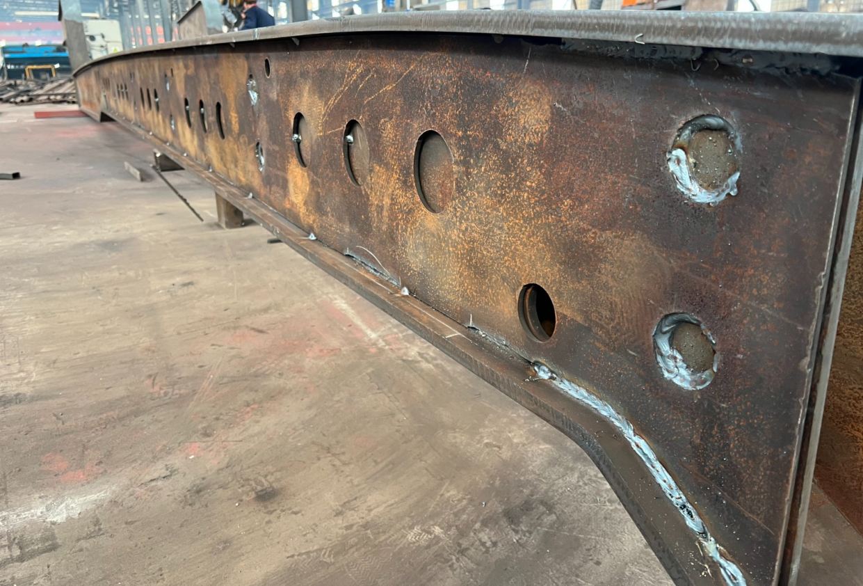 Close-up of the HUAYUE lowbed trailer's Q690 high-tensile steel main beam. Clearly detailing the thick upper and lower flanges, the robust web with engineered cutout holes, and the flawless Submerged Arc Welding structure.