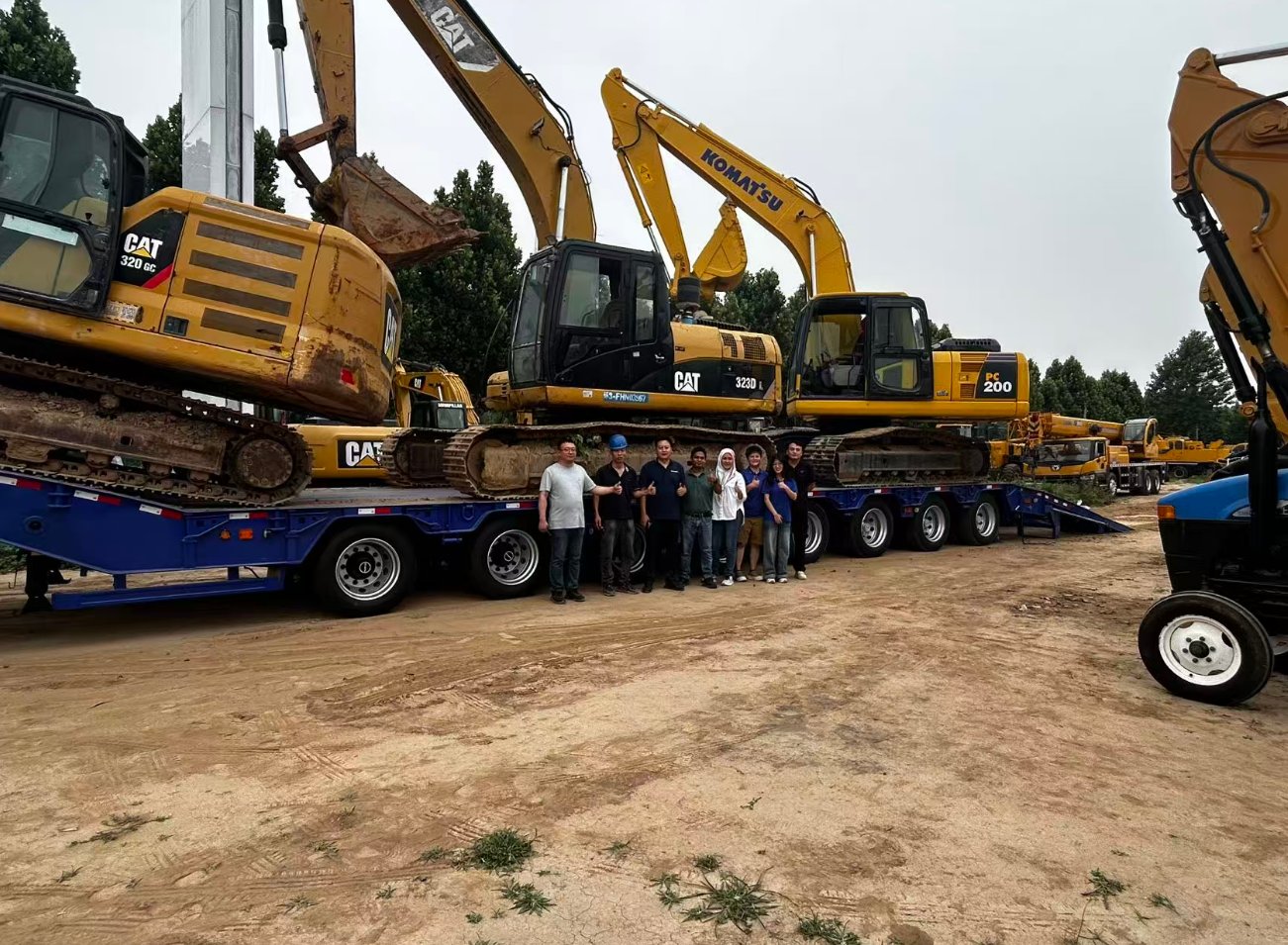 The fully loaded HUAYUE 10-axle heavy lowbed trailer holding 3 massive excavators (CAT 320 GC, CAT 323D L, and KOMATSU PC 200). The Malaysian delegation and manufacturing team celebrate the successful weight test together.