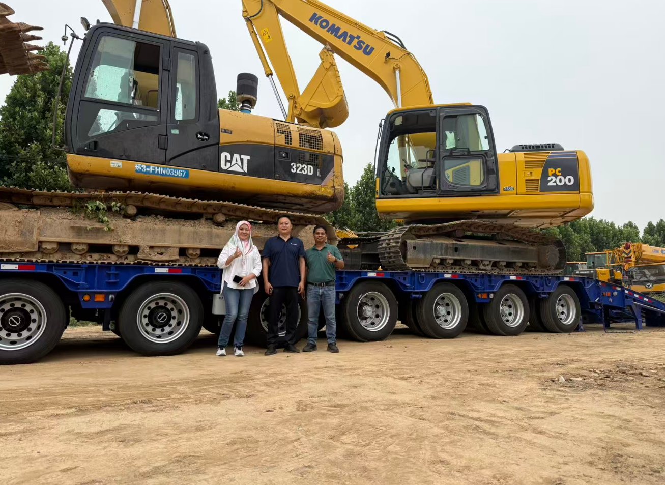Malaysian client representatives posing with HUAYUE engineers next to the 10-axle lowbed trailer. The deck is successfully loaded with a CAT 323D L and a KOMATSU PC 200 excavator, exhibiting premium structural rigidity under immense weight.