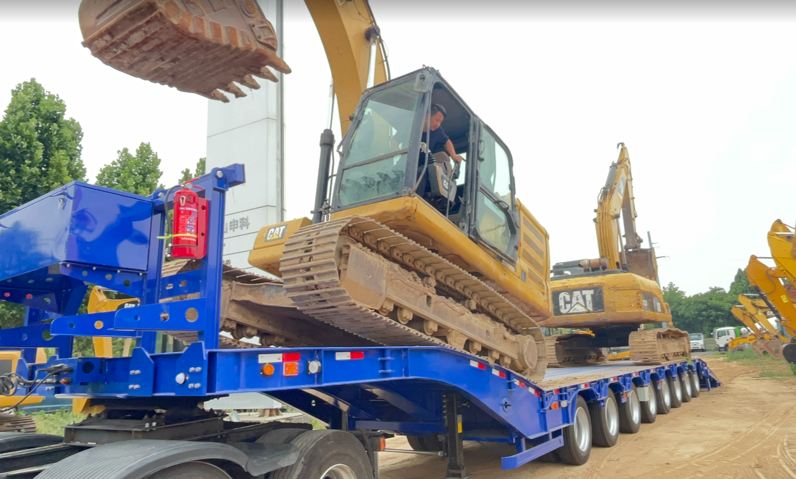 A close-up side view of an RGN trailer fully detached, with the deck flat on the ground and a heavy crawler excavator driving smoothly onto it from the front. Ensure the extreme low ground clearance is visible.