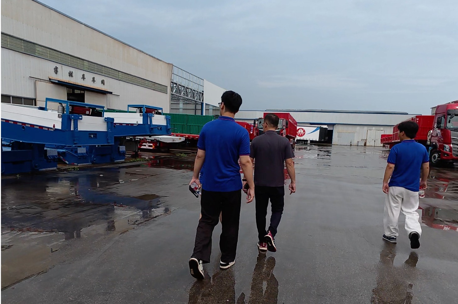 Factory visitors walking beside lowbed semi-trailer equipment in a wet yard